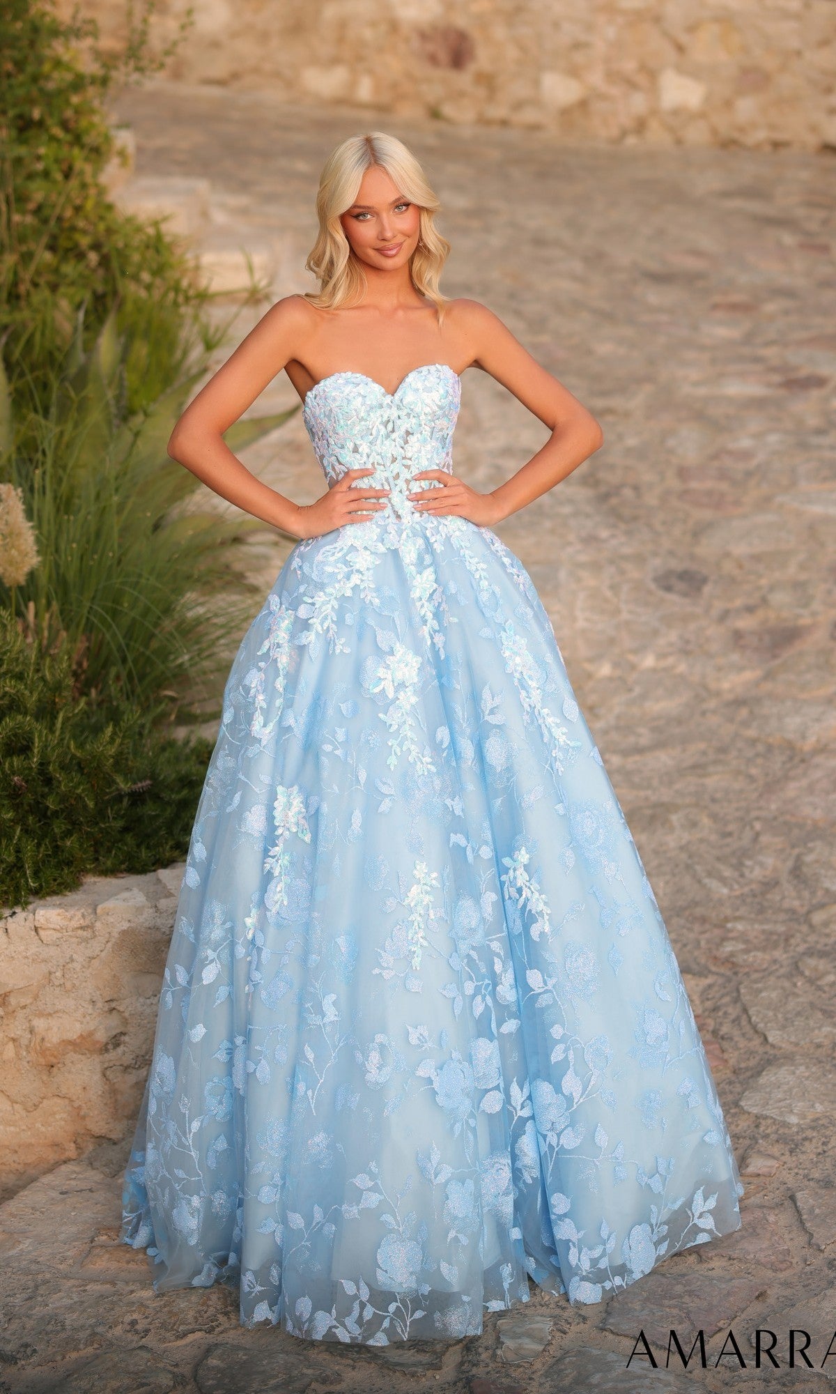 A woman stands outdoors on a stone pathway, framed by greenery and a rustic wall, wearing the Amarra 89513 long prom dress with floral-embroidered tulle and a strapless neckline.